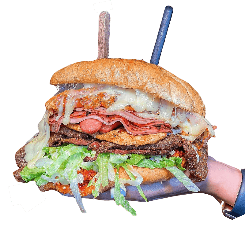 Large sandwich made with artisan bread, layers of meat, chicken, salami, melted cheese, lettuce, and sauce, held together by a hand with two toothpicks sticking through it to keep it assembled.