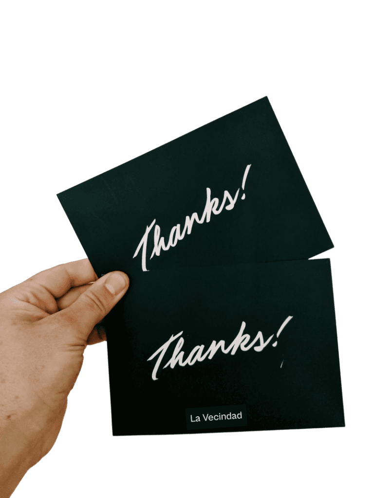 Una mano sosteniendo dos tarjetas negras con la palabra “Thanks!” en letras blancas y “La Vecindad” impreso en la parte inferior